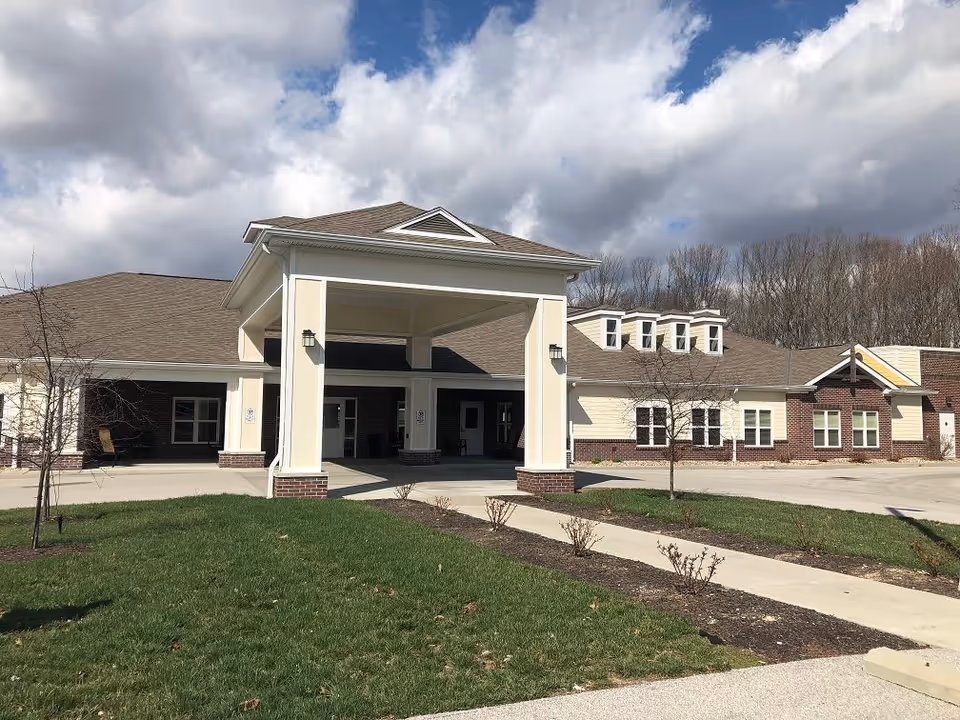 Front entrance of a single-story assisted living building with a covered drive-through portico, lawn, and cloudy sky.