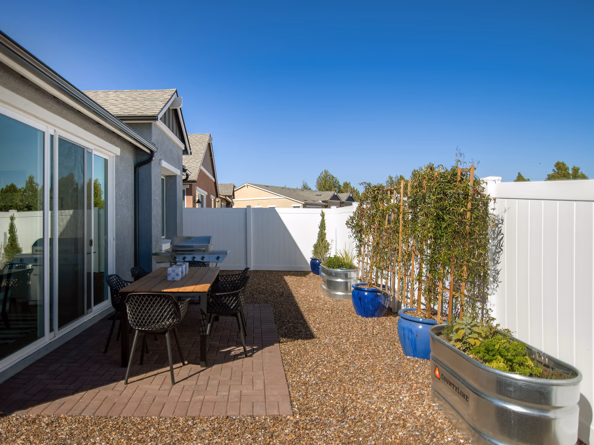 Backyard patio with a dining table and chairs, a grill, and potted plants along a white fence.