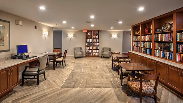 Well-lit communal library and seating area with bookshelves, tables, chairs, and a computer workstation.