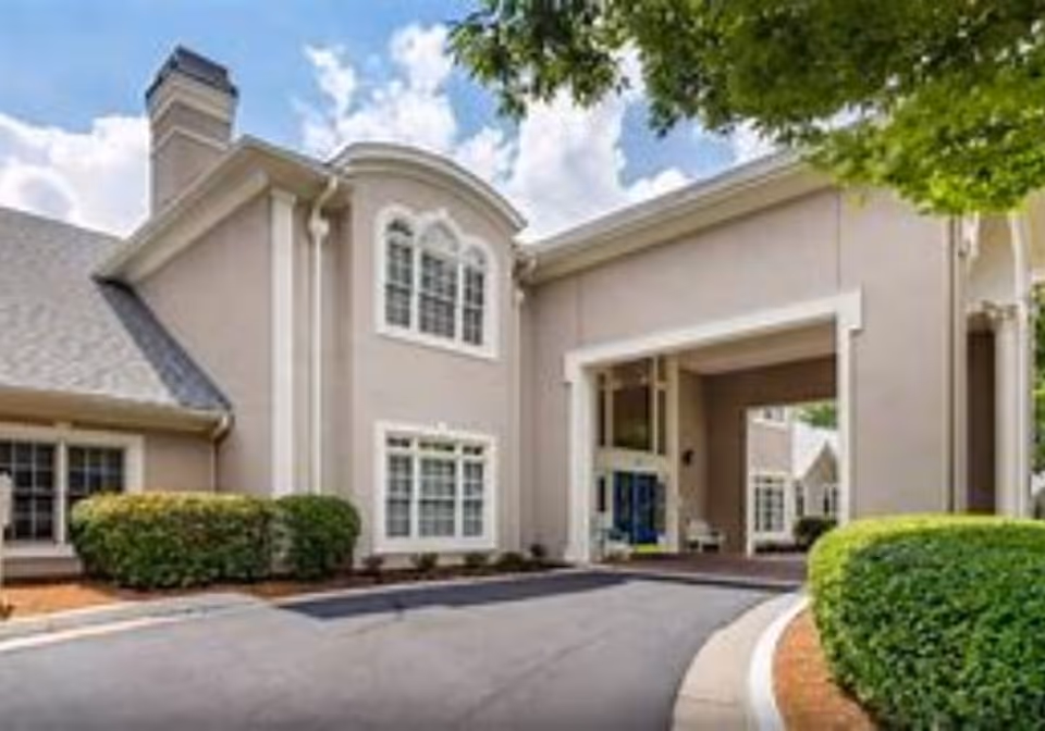 Exterior view of a senior living facility building with beige walls, white trim, multiple windows, a covered entrance, and neatly trimmed bushes along the driveway under a partly cloudy sky.