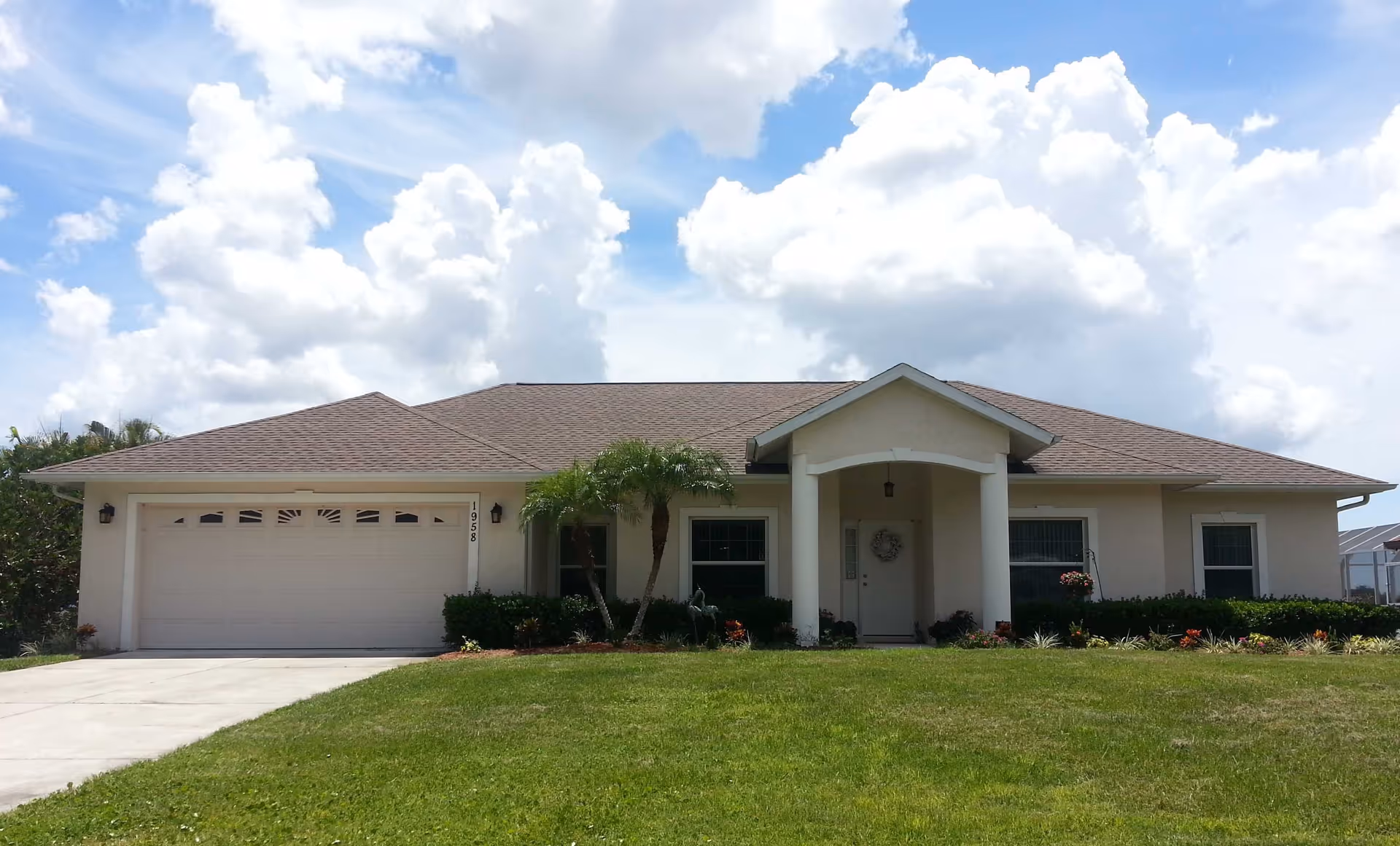 Single-story ranch-style house front with an attached garage, manicured lawn, small palm trees and a cloudy blue sky.