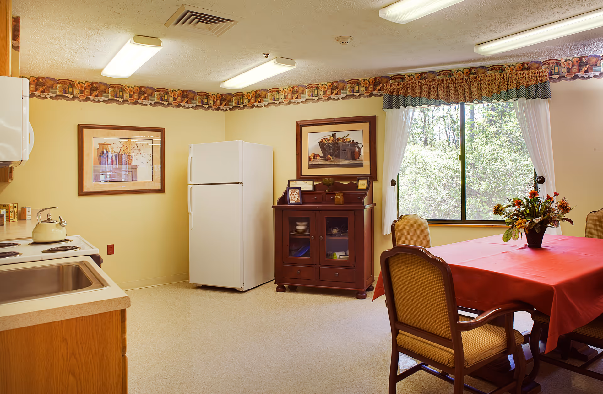 A cozy kitchen and dining area with a white refrigerator, wooden cabinets, a stove with a kettle, and a sink. There is a wooden cabinet with glass doors holding dishes, two framed pictures on the walls, and a window with curtains letting in natural light. A dining table covered with a red tablecloth is surrounded by four cushioned chairs, and a vase with flowers is placed on the table.