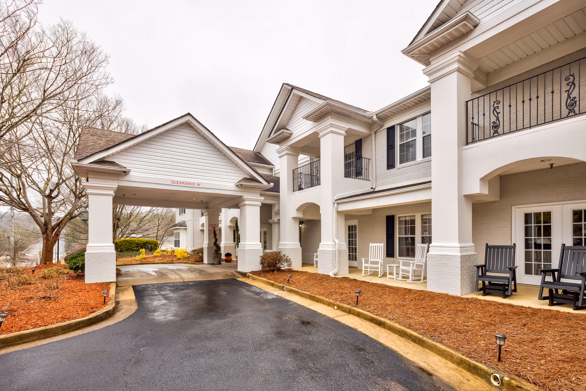 Exterior view of a senior living facility entrance with a covered driveway supported by white columns. The building has white brick walls, black window shutters, and a balcony with decorative railings. There are rocking chairs on the porch area and landscaped beds with mulch and small shrubs along the driveway.