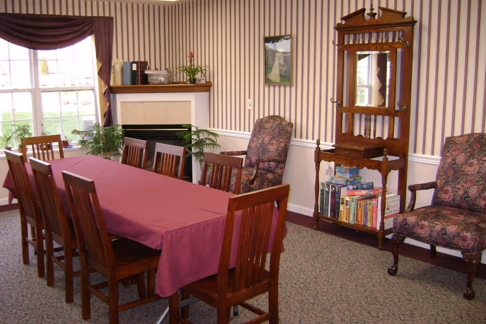 A dining room with a long wooden table covered by a maroon tablecloth surrounded by wooden chairs. The room has striped wallpaper, a fireplace with plants on the mantel, two floral upholstered armchairs, a wooden hall tree with a mirror and shelves holding books and board games, and a large window with curtains letting in natural light.