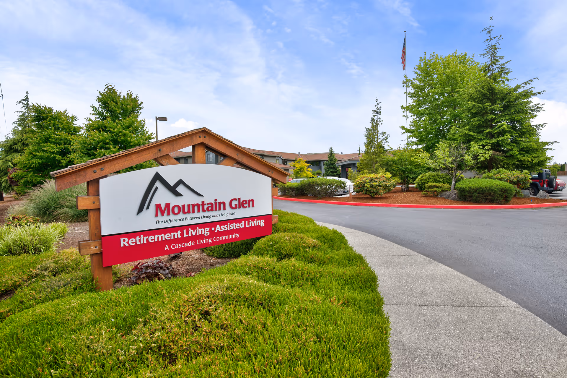 Entrance sign for Mountain Glen Retirement Living and Assisted Living community surrounded by green bushes and trees, with a paved driveway and building in the background under a partly cloudy sky.