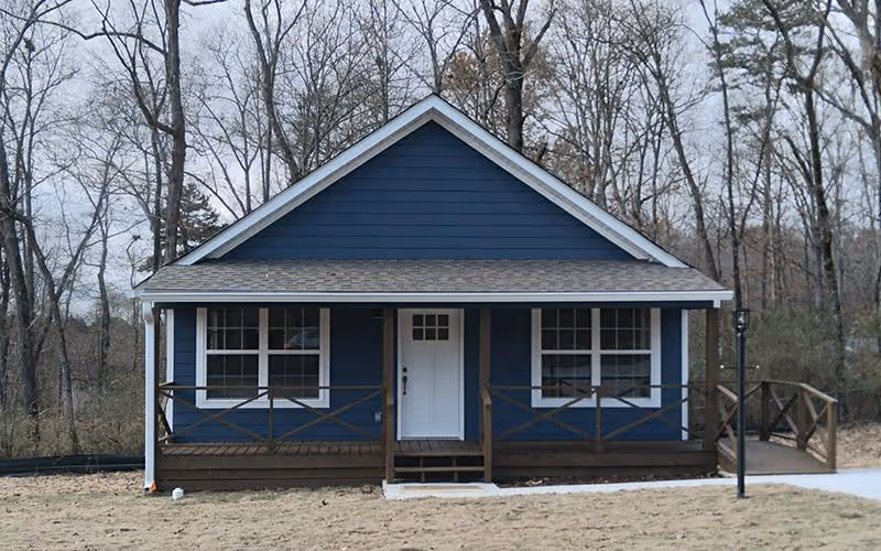 Front exterior view of a small blue house with white trim, a white front door, two windows on either side of the door, a wooden porch with railings, and leafless trees in the background.