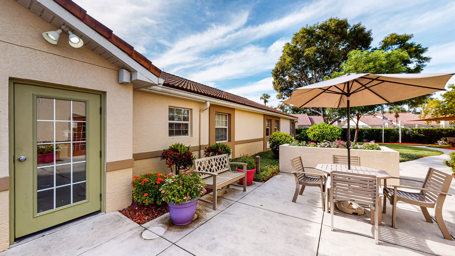 Outdoor patio area at a senior living community with a beige building featuring a green door and windows. The patio has a wooden bench, potted plants, a round table with four chairs, and a large beige umbrella. Trees and a well-maintained lawn are visible in the background under a partly cloudy sky.