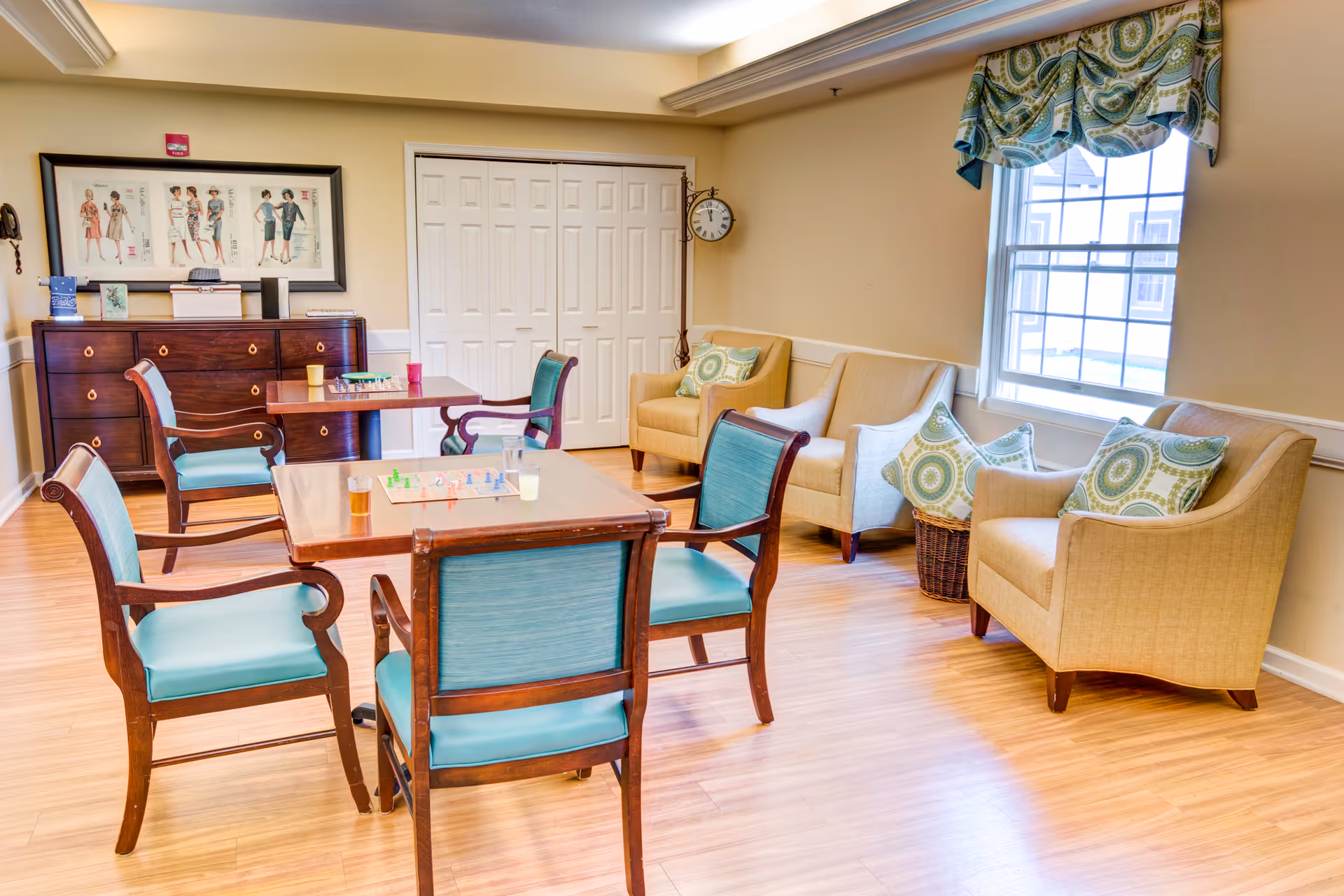 A bright and cozy common room with wooden flooring, featuring two square tables with four wooden chairs upholstered in teal fabric. On the tables are board games and drinks. Against the wall is a wooden sideboard with framed artwork above it. There are three beige armchairs with patterned cushions near a window with a decorative valance. A clock hangs on the wall near white double doors.