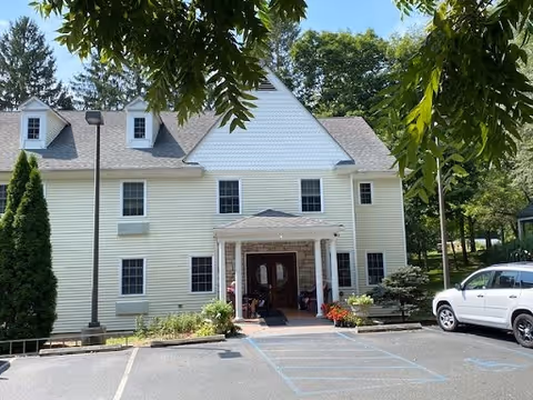 Exterior view of a two-story light yellow building with white trim and a peaked roof, surrounded by trees and greenery. There is a covered entrance with columns and a parking area in front, including a white SUV parked on the right side.