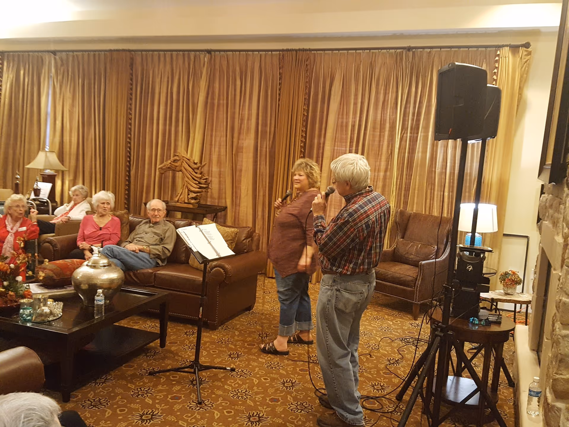 A group of elderly people seated on leather sofas in a living room with brown curtains and carpet. Two people, a man and a woman, are standing and holding microphones, possibly singing or speaking. There is a music stand with sheet music in front of the seated group, and a speaker on a stand to the right.