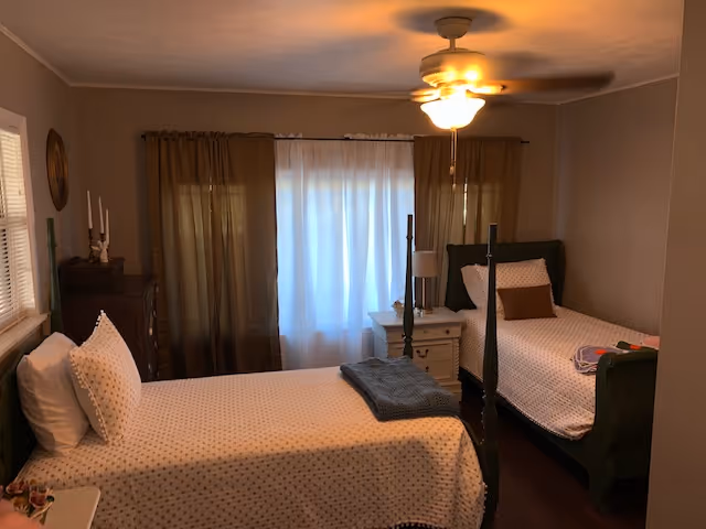 A cozy bedroom with two single beds, each with white polka dot bedding and pillows. There is a ceiling fan with a light fixture above, a window with sheer white curtains and brown drapes, a white nightstand with a lamp between the beds, and a wooden dresser with candles on top in the corner.