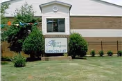 Exterior view of a building with a sign in front that reads 'Senior Villages' along with a phone number. The building has a beige and white facade with a window and some bushes and trees around the sign.