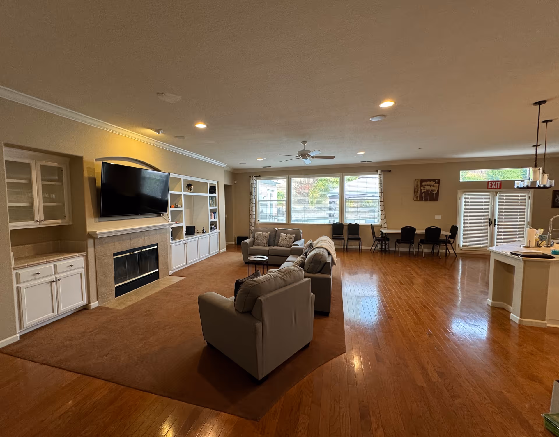 A spacious living room area with wooden floors and a large brown rug. The room features a fireplace with a mounted flat-screen TV above it, built-in white cabinets and shelves, and several gray upholstered chairs and a sofa arranged around a small round table. Large windows with blinds let in natural light, and there is a dining area with tables and chairs near glass doors with an exit sign above. A kitchen island with a sink and hanging light fixtures is visible on the right side.