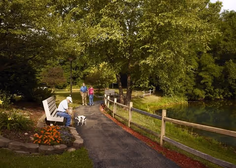 A peaceful outdoor walking path lined with a wooden fence and surrounded by lush green trees and plants. A man sits on a bench petting a small dog, while a couple walks along the path in the background near a pond.
