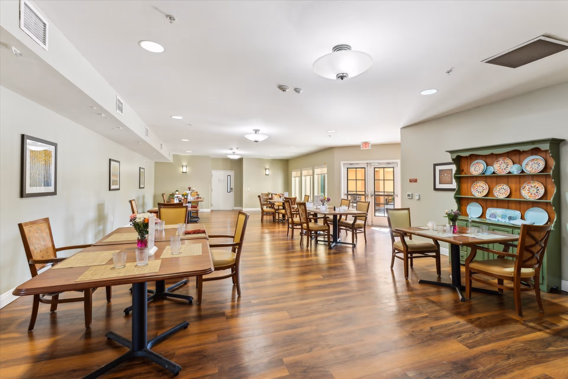 A bright dining room with wooden floors and multiple wooden tables and chairs arranged neatly. Each table has placemats, glasses, and small flower arrangements. The walls are light-colored with framed artwork, and there is a green hutch displaying decorative plates. Large windows and glass doors allow natural light to enter the room.