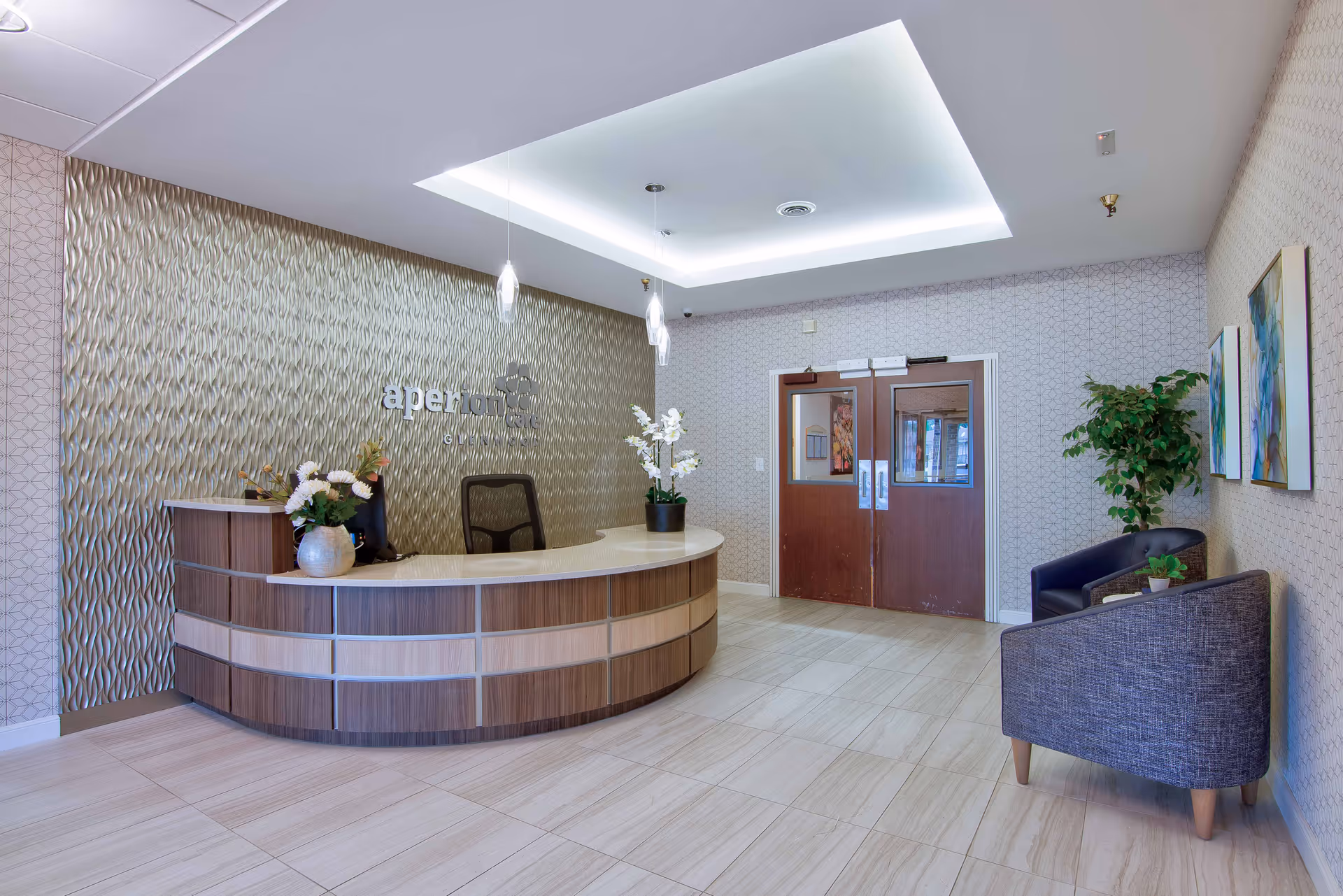 Reception area with a curved wooden desk, a black office chair, decorative plants and flowers on the desk, textured wall behind the desk with the word 'aperture' displayed, two wooden double doors, two gray armchairs with a small table and plant between them, and framed artwork on the wall.