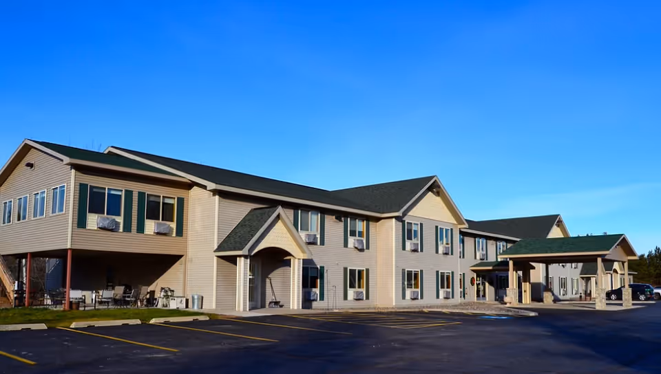 Exterior view of Cedar Hill Assisted Living & Senior Housing building under a clear blue sky, showing a two-story structure with beige siding, green shutters, multiple windows with air conditioning units, a covered entrance, and an empty parking lot in front.