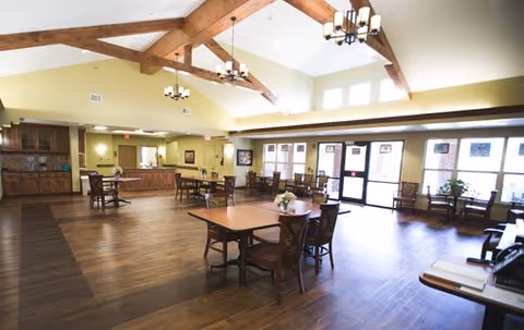 Spacious dining room with wooden floors, multiple tables and chairs, large windows letting in natural light, exposed wooden ceiling beams, and chandeliers. There is a counter area with cabinets along one wall and some framed artwork on the walls.
