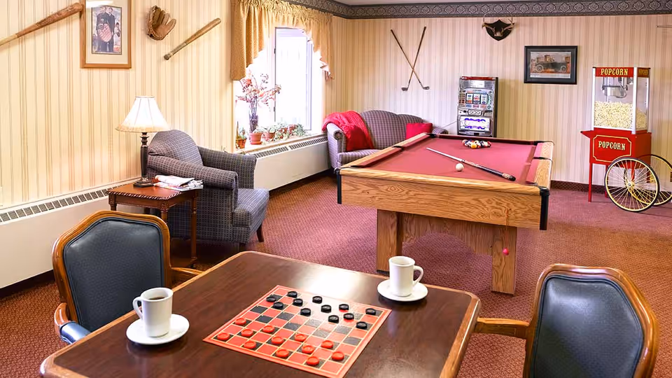 A cozy recreational room with a pool table, a checkers board on a wooden table with two chairs, two armchairs near a window with plants, a popcorn machine, and wall decorations including golf clubs and framed pictures.