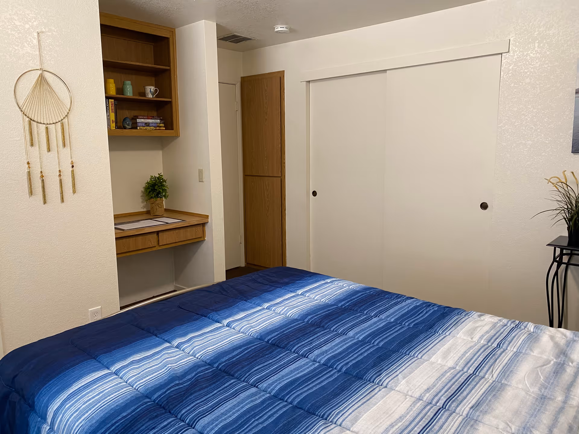A bedroom with a bed covered in a blue and white striped comforter. There is a built-in wooden desk with shelves above it holding books and decorative items. A small potted plant is on the desk. The room has white walls, a wooden door, and a white sliding closet door. A decorative hanging piece is on the left wall and a small table with a plant is on the right side.