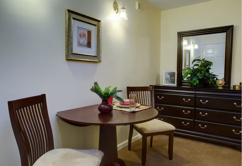 A small dining area with a round wooden table set for two, featuring a red vase with green foliage, two wooden chairs with beige cushions, a large dark wooden dresser with a mirror above it, and a framed picture on the wall under a wall-mounted light fixture.