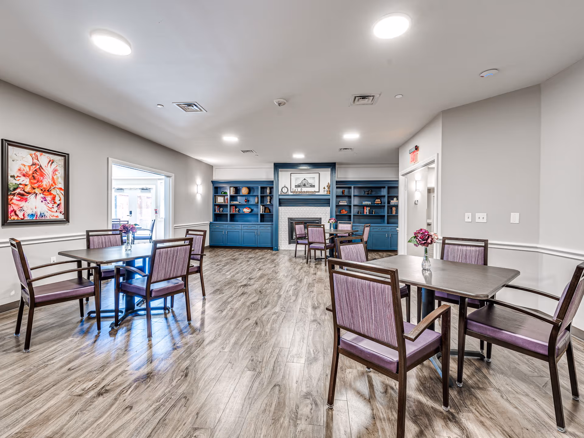 A bright and spacious common area with wooden flooring, featuring several tables with purple cushioned chairs. The room has a blue built-in bookshelf and a white fireplace at the far end. There is a colorful floral painting on the left wall and small vases with flowers on the tables. The ceiling has recessed lighting and the walls are painted light gray.