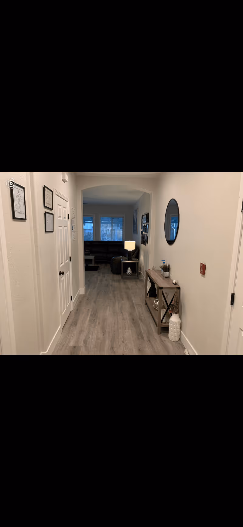A hallway with light-colored walls and wood flooring leading to a living room area with a dark sofa and a lit table lamp. On the right side of the hallway, there is a console table with decorative items and a round mirror above it. On the left side, there are framed documents or pictures on the wall and a closed white door.