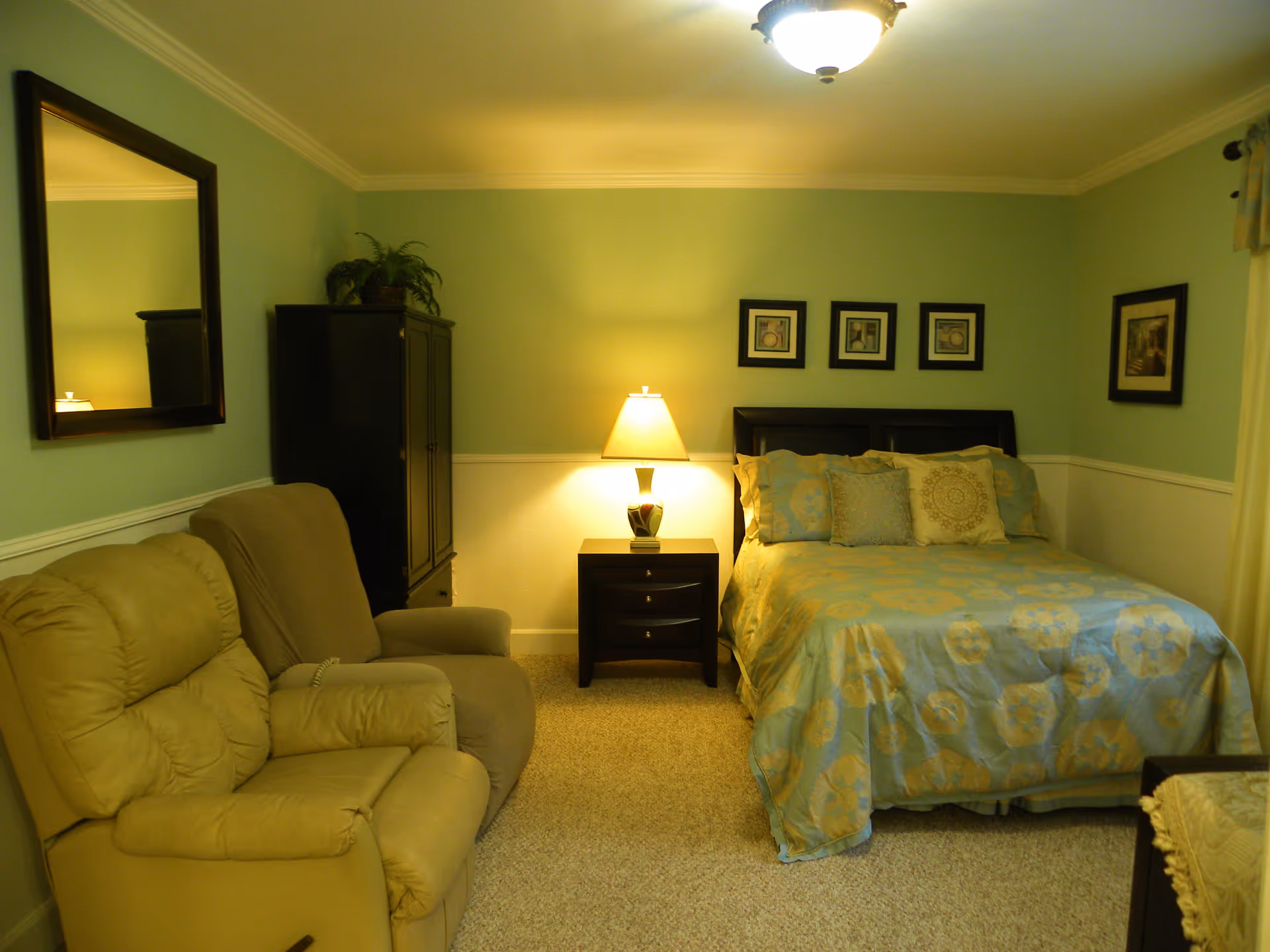 A cozy bedroom with a bed covered in a blue and yellow floral bedspread, several pillows, a nightstand with a lamp, two armchairs, a black cabinet with a plant on top, a large mirror on the wall, and framed pictures hanging above the bed and on the adjacent wall.