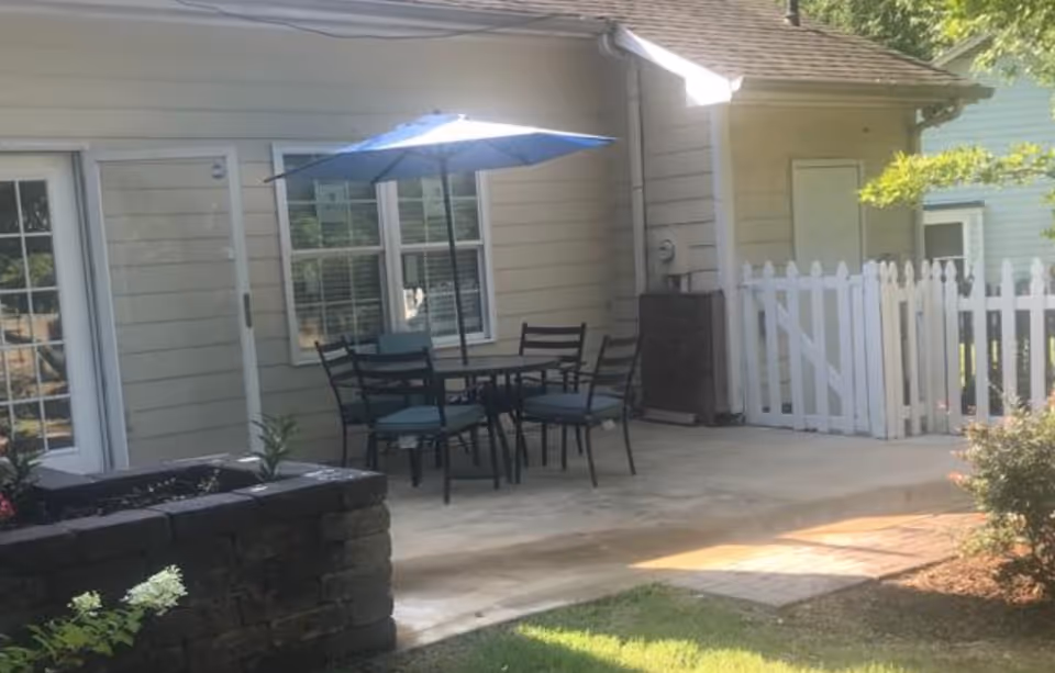 Small outdoor patio with a round table and chairs under a blue umbrella next to a light-colored building and white picket fence.