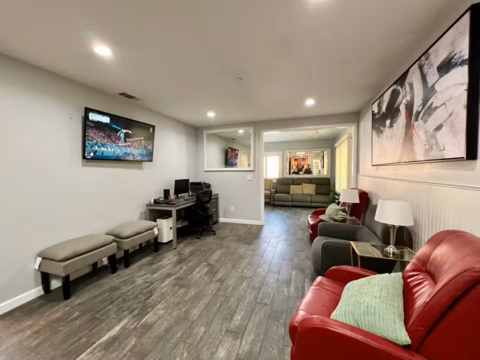 A cozy living room area with a red leather armchair and a gray armchair, both with pillows, and a side table with a lamp. The room has wood-look flooring and light gray walls. A large abstract painting hangs on the right wall. In the background, there is another seating area with a green sofa, and a wall-mounted TV is visible on the left wall above a small desk with a computer and office chair. The room is well-lit with recessed ceiling lights.