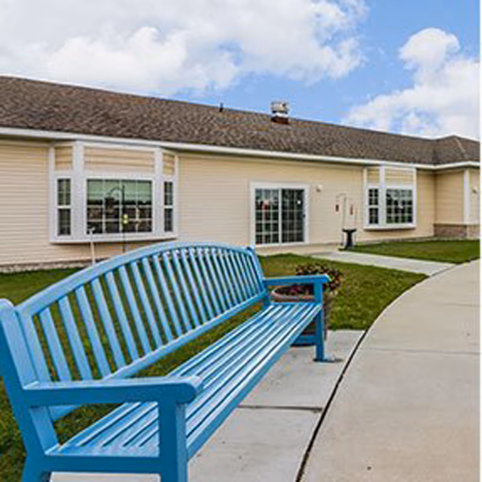 A bright blue outdoor bench on a sidewalk in front of a single-story beige building with windows, lawn, and a partly cloudy sky.