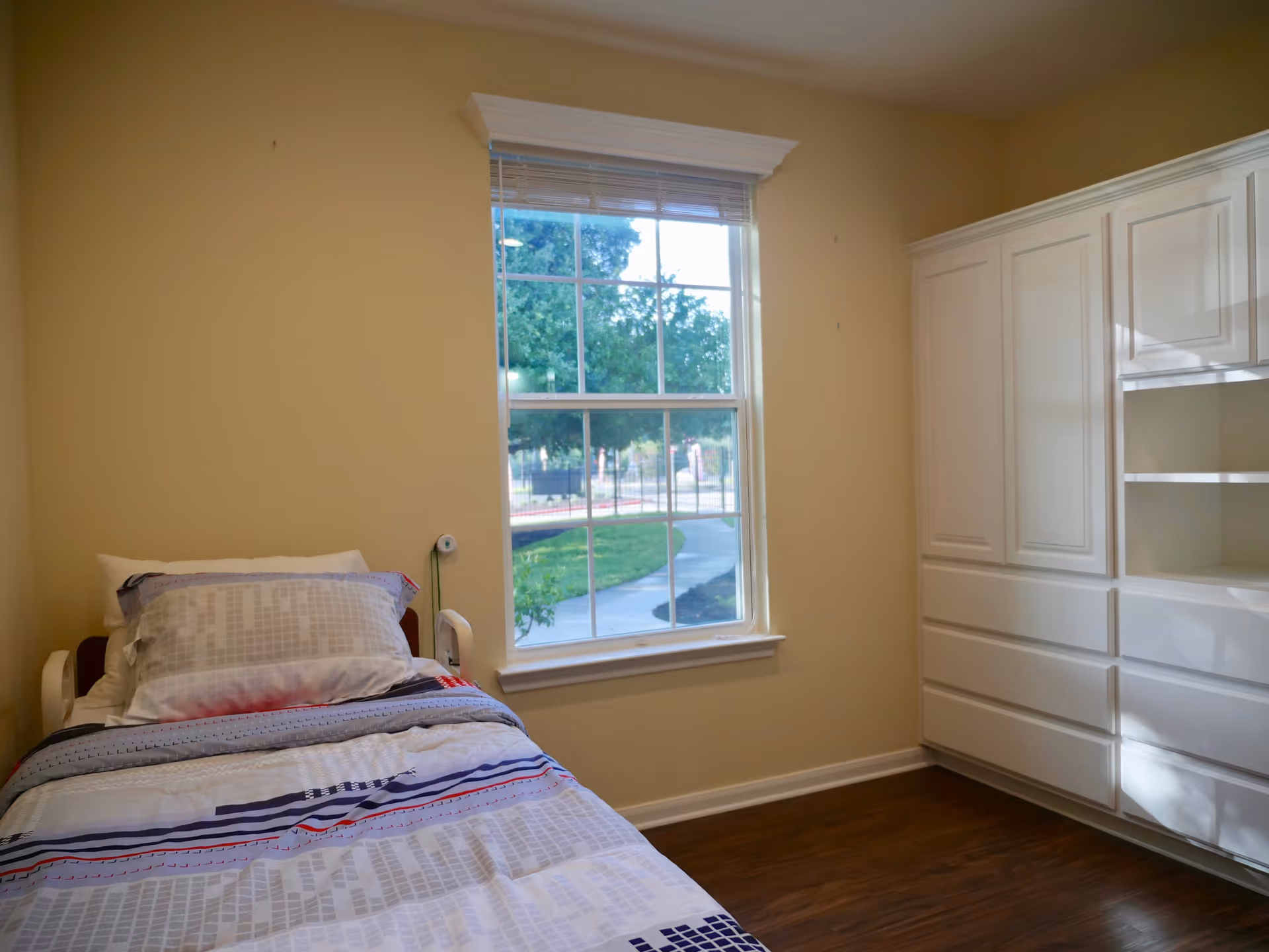 A simple bedroom with a single bed made up with patterned bedding and pillows. There is a large window with a view of a sidewalk and greenery outside. On the right side of the room, there is a white built-in cabinet with multiple drawers and shelves. The walls are painted beige and the floor is wooden.