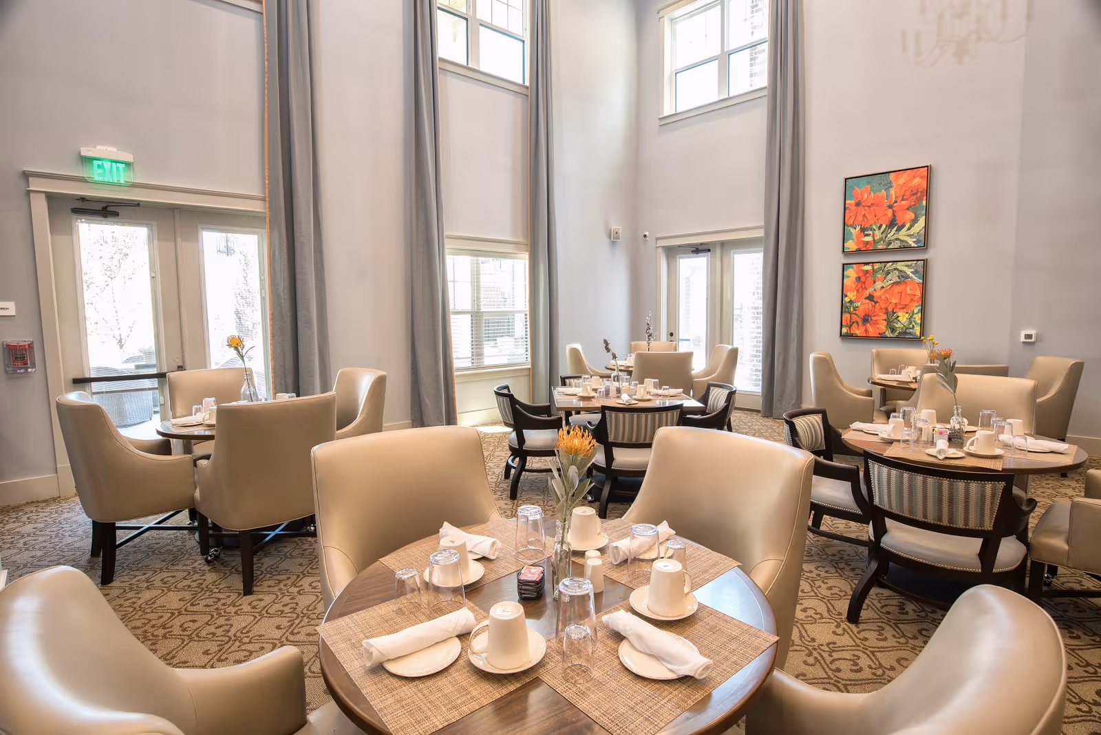 A bright dining room with several round tables set with white cups, plates, napkins, and glasses. The room features high ceilings, large windows with gray curtains, beige upholstered chairs, and patterned carpet. Two colorful floral paintings hang on the wall.