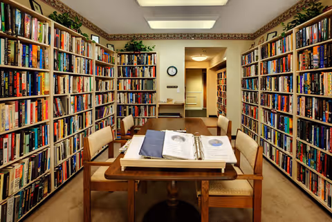 A cozy library room with tall bookshelves filled with books lining both sides of the walls. In the center, there is a wooden table with four cushioned chairs around it. On the table, there is an open binder with photos and documents. The room is well-lit with ceiling lights and decorated with some green plants on top of the bookshelves.