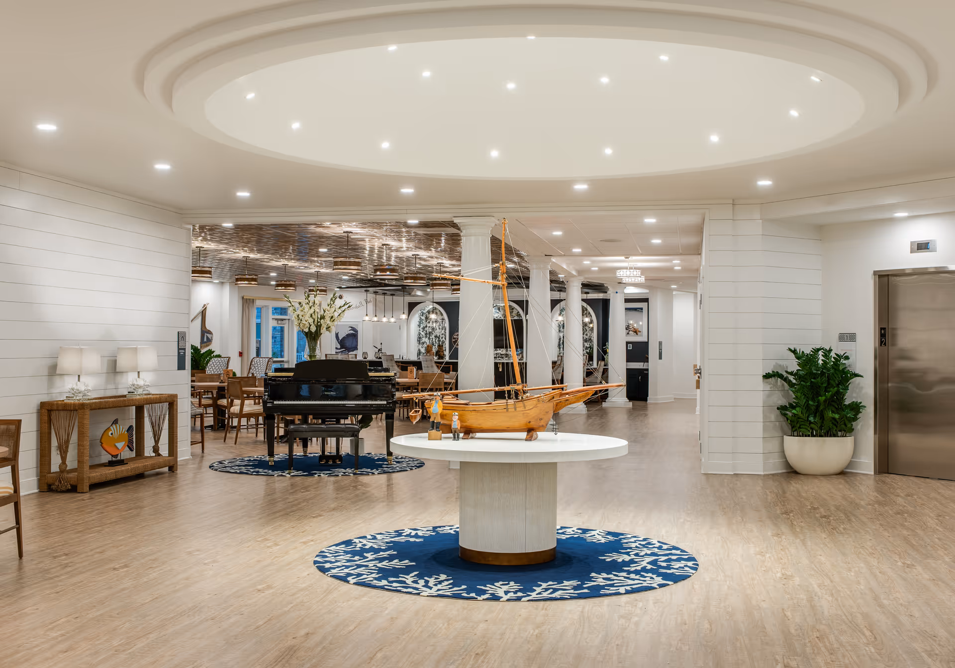 A spacious and well-lit common area in a senior living facility featuring a round table with a wooden model sailboat on a blue rug with white coral patterns. In the background, there is a black grand piano on a similar rug, several dining tables and chairs, white columns, and decorative wall art. The floor is light wood, and there is an elevator on the right side with a large potted plant nearby.