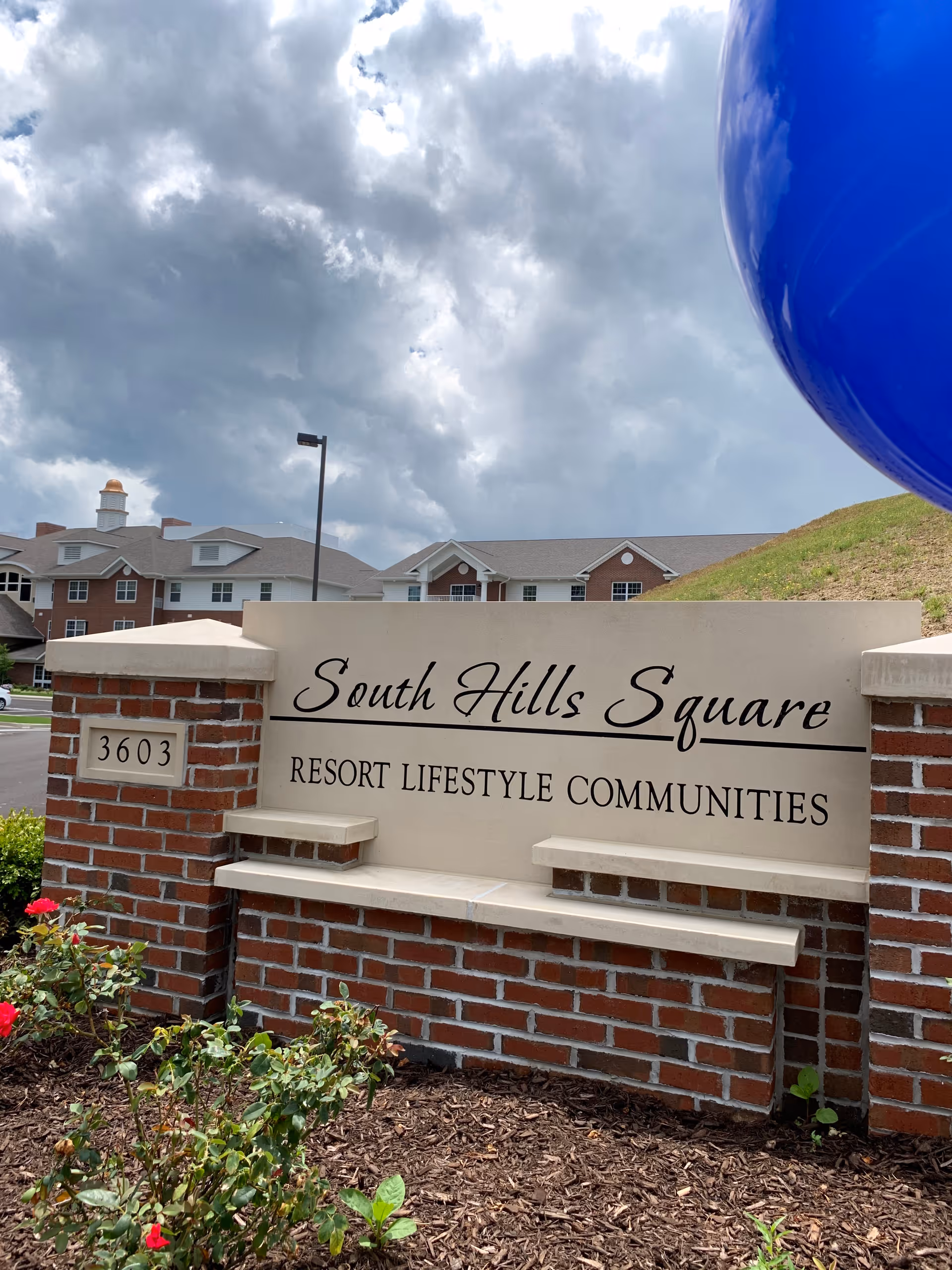 Entrance sign for South Hills Square Resort Lifestyle Communities made of brick and stone with flowers and greenery in front, residential buildings and a cloudy sky in the background.