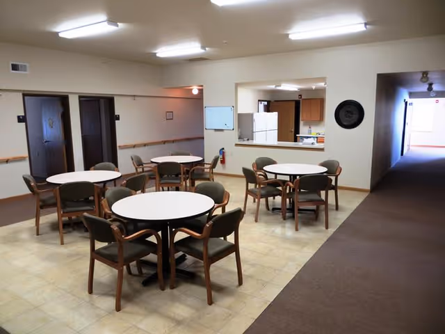 A common area in an assisted living facility with four round tables, each surrounded by four chairs. The room has beige walls and a tiled floor area with a carpeted hallway leading to other rooms. There is a kitchen pass-through window with a refrigerator visible, and several doors along the walls.