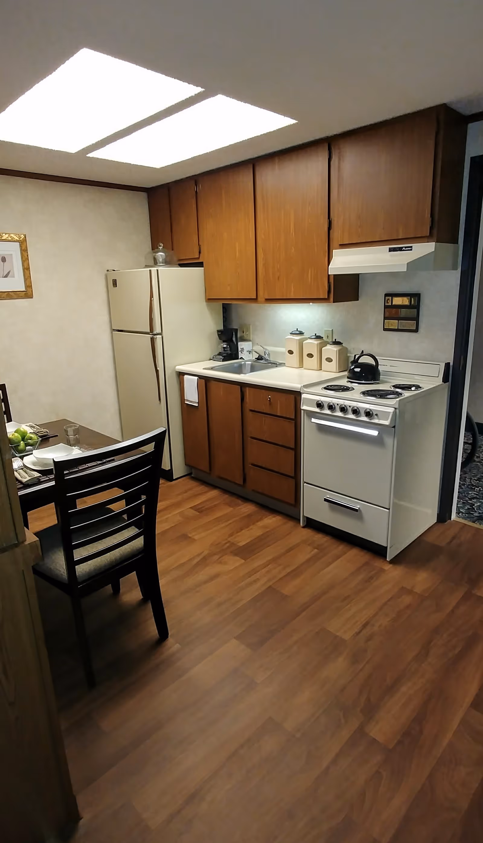 A small kitchen area with wooden cabinets, a white refrigerator, a white stove with a kettle on top, a sink, and a dining table with two chairs. The floor is wooden, and there are two large ceiling lights.