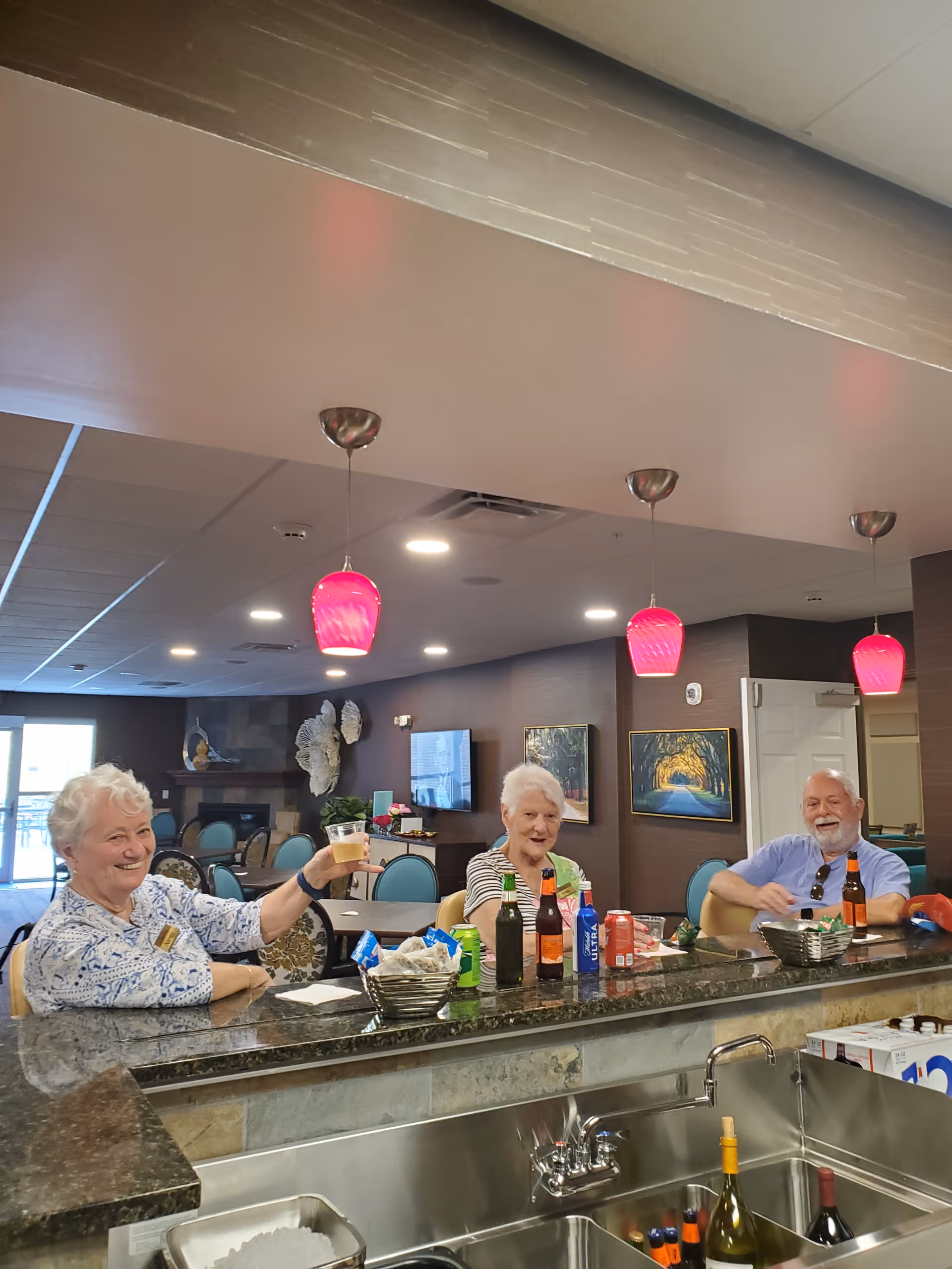 Three elderly people sitting at a bar counter in a retirement community, smiling and enjoying drinks. The setting includes a granite countertop, hanging pink pendant lights, and a cozy interior with paintings on the wall and a TV in the background.