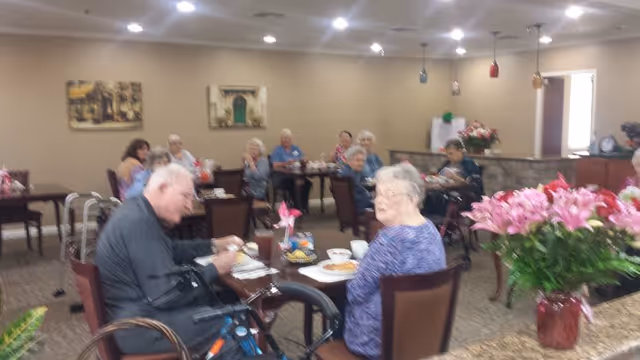 A group of elderly people sitting at tables in a dining area, eating and socializing. The room has beige walls, ceiling lights, and decorative paintings on the walls. There is a large bouquet of pink flowers on a counter in the foreground.