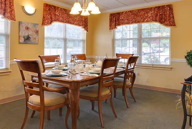 A dining room with a wooden table set for six people, including plates, glasses, and silverware. The room has large windows with red patterned valances, beige walls, and a carpeted floor. A chandelier with five lights hangs above the table, and there is a floral painting on the wall.