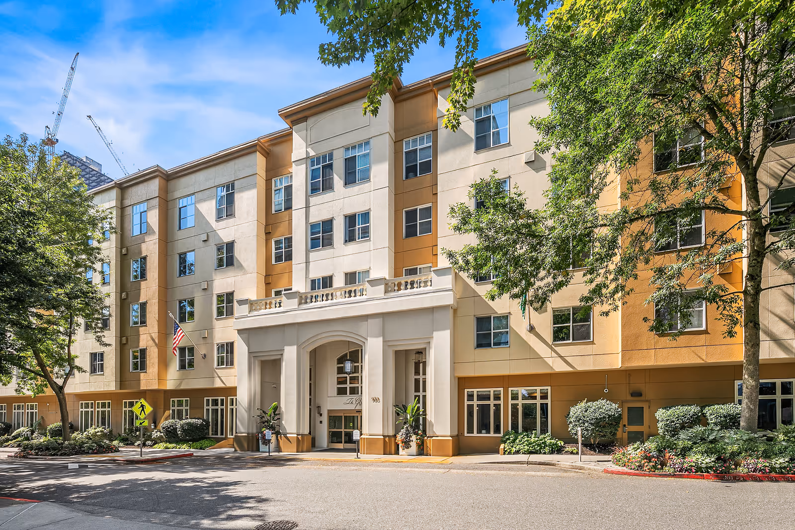 Exterior view of a multi-story senior living facility building with beige and light brown walls, multiple windows, an arched entrance, and surrounding trees and landscaping under a blue sky.