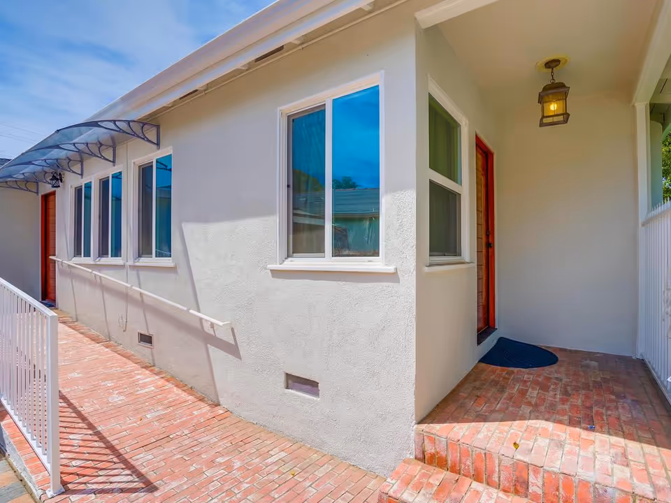 Brick walkway and ramp leading to the side entrance of a single-story light-colored building with several windows and a covered doorway.