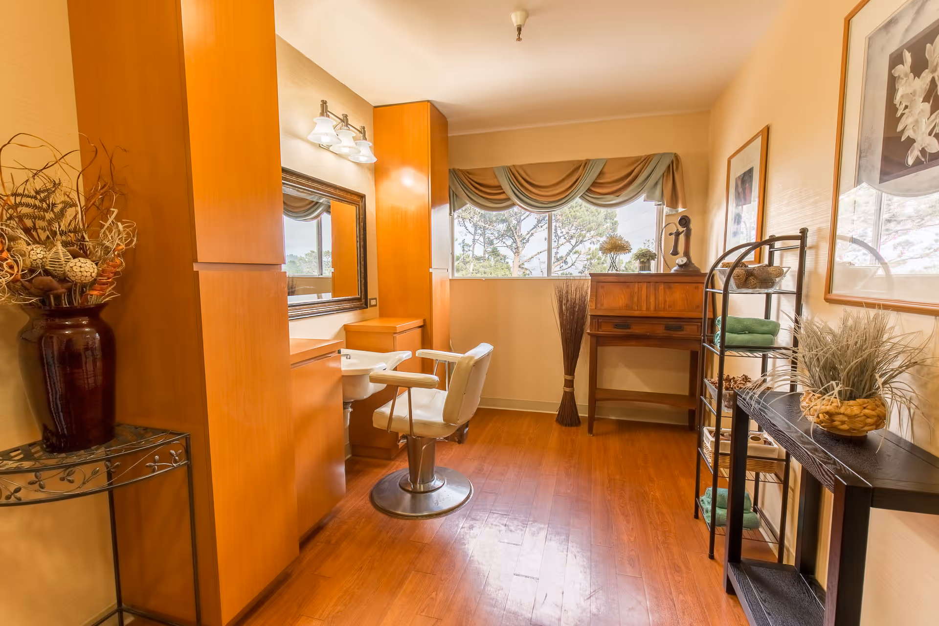 A well-lit room with wooden flooring and light-colored walls featuring a vanity area with a mirror, a white swivel chair, and built-in wooden cabinets. There is a window with a decorative valance letting in natural light. The room also contains a wooden desk with decorative items, a metal shelving unit with green towels and other decor, and a small table with a large vase of dried plants.