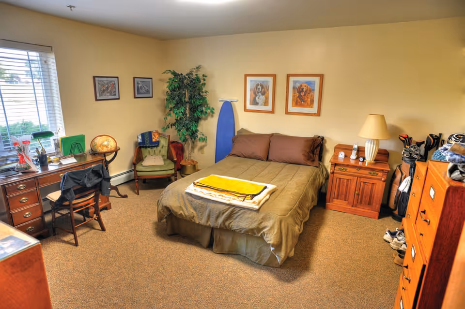 Furnished bedroom in a senior living facility with bed, desk, dresser, and decor.