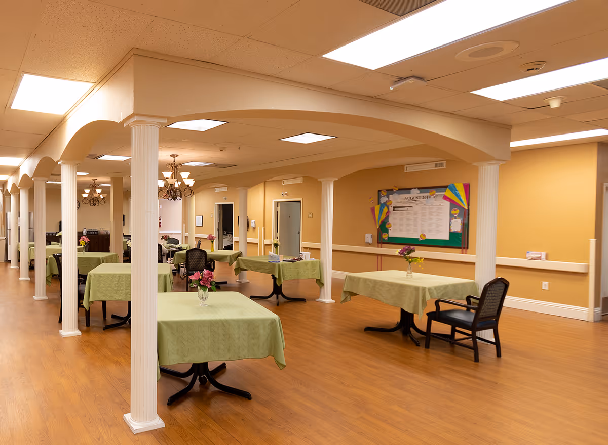 Interior view of a dining room in a senior living facility with multiple tables covered in green tablecloths, each adorned with a small vase of flowers. The room features wooden flooring, white columns, beige walls, and ceiling lights. A bulletin board with colorful decorations is visible on one wall.