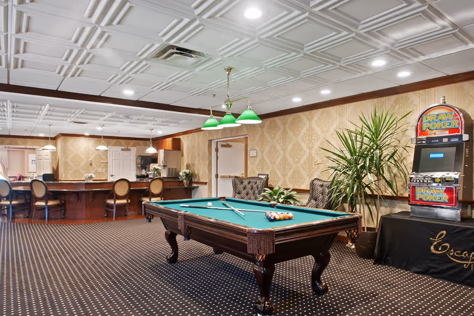 A recreational room featuring a pool table with billiard balls and cues, a slot machine labeled 'Draw Poker,' several chairs, a long counter with bar stools, and decorative plants. The room has patterned wallpaper, a coffered ceiling with recessed lighting, and a carpeted floor with a dotted pattern.