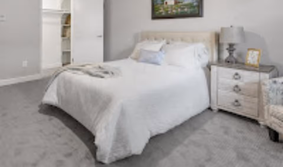 A neatly made bed with white bedding in a bright bedroom, a nightstand with lamp and framed photo to the right and a closet doorway in the background.