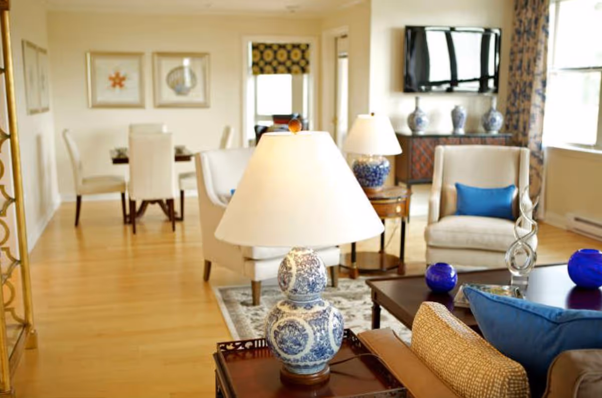 A bright and cozy living room with beige armchairs, blue accent pillows, a wooden coffee table with decorative items, and a blue and white table lamp in the foreground. In the background, there is a dining area with a table and chairs, framed artwork on the wall, and a flat-screen TV mounted on the wall above a cabinet with blue and white vases.