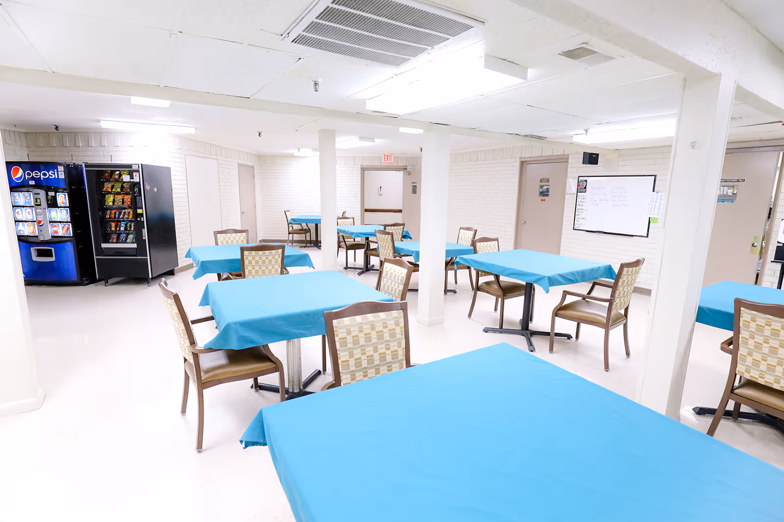 A bright dining area with multiple tables covered in blue tablecloths and surrounded by chairs. In the background, there are vending machines against a white brick wall and a whiteboard mounted on the wall. The room has white walls and ceiling with fluorescent lighting.